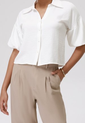 Femme portant un chemisier blanc à boutons avec des manches bouffantes et un pantalon beige taille haute, main dans la poche, bracelet en or au poignet.