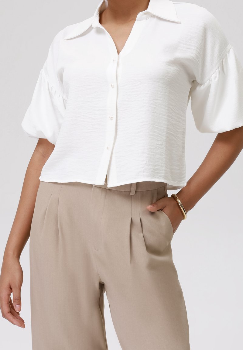 Femme portant un chemisier blanc à boutons avec des manches bouffantes et un pantalon beige taille haute, main dans la poche, bracelet en or au poignet.