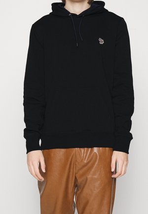 Person trägt schwarzen Hoodie mit kleinem buntem Zebra-Logo auf der Brust und braune Lederhose, steht vor einfarbigem Hintergrund.