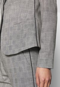 Gros plan sur une personne portant un costume gris à carreaux avec des détails de passepoil noir, montrant la poche de la veste et la manche, et sa main détendue le long du corps.