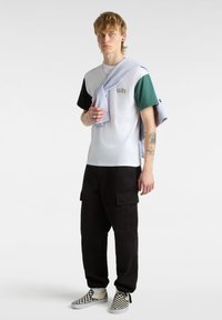 Weißes T-Shirt mit schwarzen und grünen Ärmeln, mit "VANS"-Logo. Schwarze Cargo-Hosen und schwarz-weiß karierte Slip-On-Schuhe. Hellblauer Pullover über die Schultern drapiert.