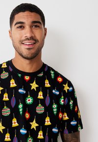 Sort kortærmet t-shirt med farverige julepyntsmønstre, herunder klokker, stjerner og kugler i forskellige former.