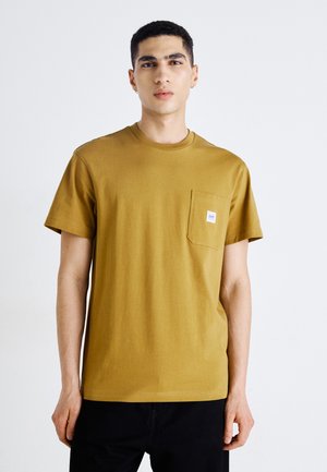 Camiseta de algodón mostaza con cuello redondo, mangas cortas y un bolsillo en el pecho con una pequeña etiqueta de la marca. Textura suave.