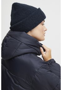 Berretto a maglia blu navy con finitura testurizzata; indossato con un giubbotto imbottito scuro con colletto alto. Inquadratura ravvicinata del profilo laterale.