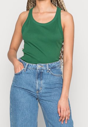 Vrouw met gevlochten haar, gekleed in een groen tanktop en hoge blauwe spijkerbroek, één hand in haar zak en de andere ontspannen langs haar zijde.