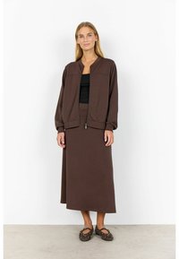 Frau in brauner Kapuzenjacke und passendem langen Rock mit schwarzem Oberteil und mit Nieten besetzten braunen Ballerinas, die auf einem Betonboden steht.
