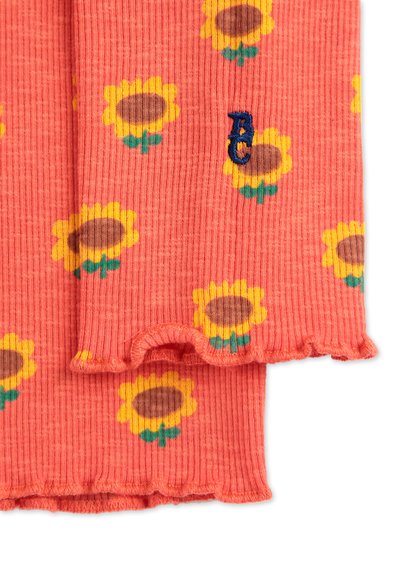 Bufanda de tela acanalada en color coral con un patrón de girasoles amarillos y acentos verdes. Presenta un borde ondulado y un pequeño logo bordado en azul.