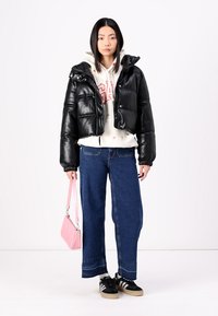 Zwarte cropped pufferjack met een glanzende afwerking, witte hoodie met rode letters, blauwe wijde jeans en een roze handtas. Zwarte sneakers maken de look compleet.