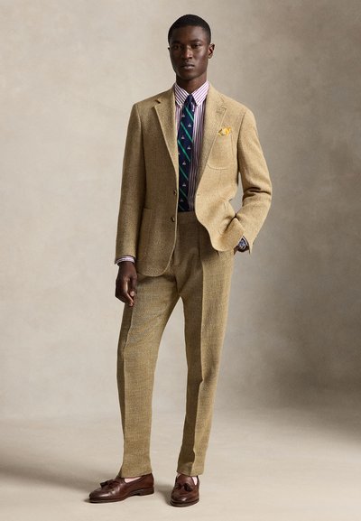 Homme portant un costume beige texturé avec une chemise à rayures, une cravate bleu marine à motifs, un mouchoir de poche jaune et des mocassins à glands marron, debout avec une main dans la poche.