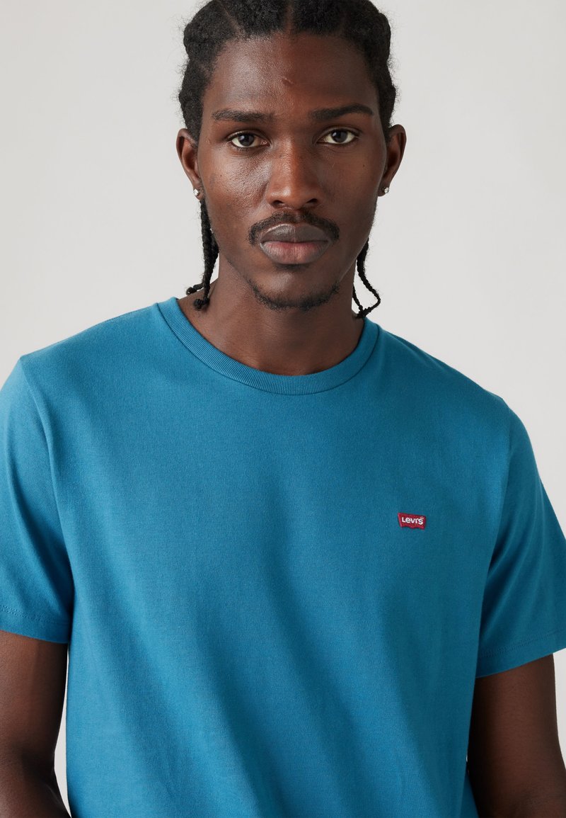 Hombre con el pelo trenzado y pendientes, vistiendo una camiseta azul de Levi’s, mirando hacia adelante sobre un fondo claro y liso.