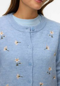Jasnoniebieski dziany kardigan z małymi, haftowanymi białymi i żółtymi kwiatami, zapinany pod kołnierzykiem, noszony na jasnoniebieskiej bluzce.