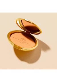 Ein runder Kompaktspiegel mit einem goldenen Rand, der ein pfirsichfarbenes Puder enthält, geprägt mit "NUXE" und einen Schatten auf einer beigen Oberfläche wirft.
