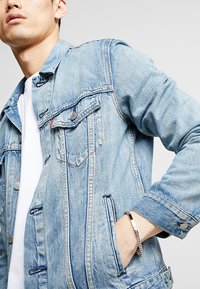 Světle modrá denimová bunda s špičatým límcem, předními kapsami a stříbrnými kovovými detaily. Má volný střih a vybledlou texturu.