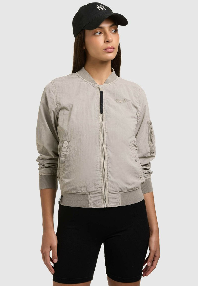 Femme portant une veste bomber beige zippée, un short de cyclisme noir et une casquette noire avec un logo blanc, regardant de côté.