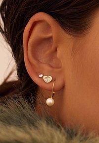 Goldene Hängerstücke mit einem Perlenanhänger, kombiniert mit herzförmigen Ohrsteckern, die mit kleinen Steinen verziert sind, und einem kleineren, diamantähnlichen Stecker.