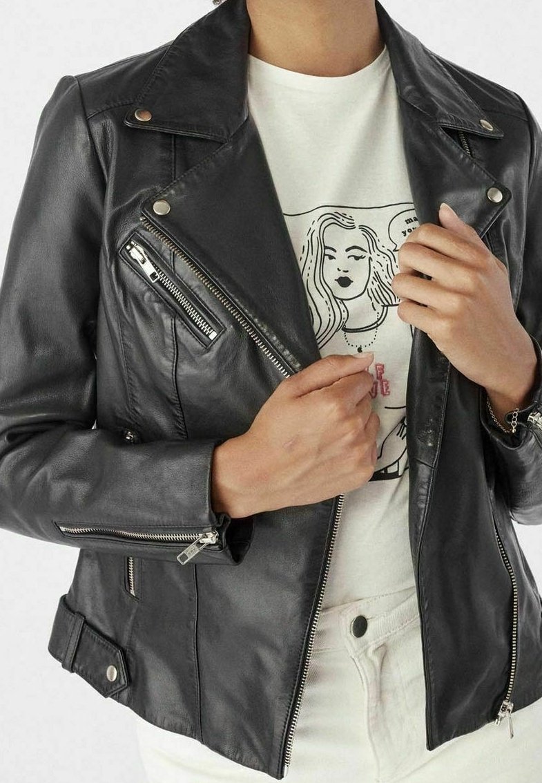 Personne portant une veste en cuir noire par-dessus un t-shirt graphique blanc et un pantalon blanc, ajustant le col de la veste avec ses deux mains.
