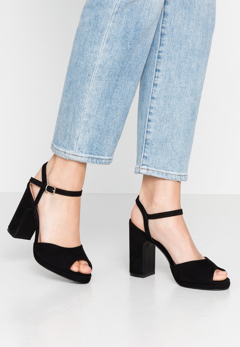 Pieds portant un jean court bleu clair et des sandales à talons blocs en suède noir avec bouts ouverts, brides à la cheville et boucles.