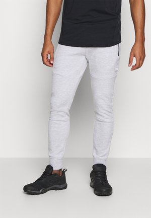 Homme portant un pantalon de jogging gris clair et des chaussures de sport noires, debout devant un fond clair uni.