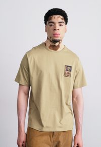 Beige katoenen t-shirt met een ronde hals, korte mouwen en een voorpatch met twee logo-ontwerpen in zwart en bruin.