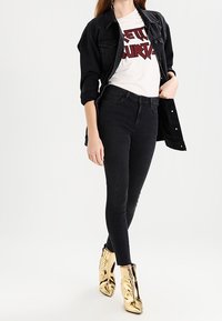 Svart jeansjacka över en vit grafisk t-shirt med röd text, matchad med svarta tajta jeans och metalliska guldankelstövlar.