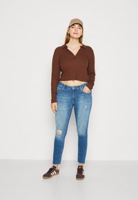 Hnědý zkrácený top s dlouhým rukávem, modré skinny džíny s vybledlými detaily a hnědé tenisky s bílými pruhy; neutrální čepice dotváří celý vzhled.