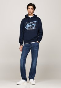 Sudadera azul marino con texto gráfico en blanco y azul claro, combinada con jeans de mezclilla azul y zapatillas blancas. Diseño minimalista y corte relajado.