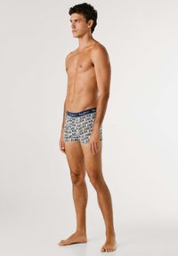 Junger Mann, der barfuß steht und bedruckte Boxershorts mit dem Text "Pepe Jeans London" trägt, vor einem schlichten, hellen Hintergrund.