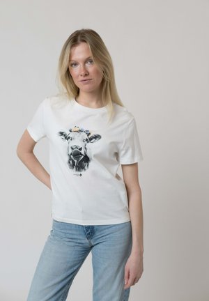 Blonde woman wearing a white t-shirt with a black and white cow face graphic adorned with blue flowers, paired with light blue jeans.