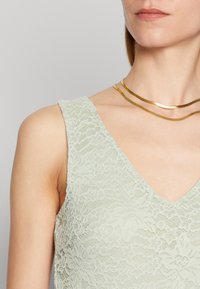 Ljusgrön spetsblus med V-ringning, blommönster och texturerat tyg, parat med gyllene lager av halsband.