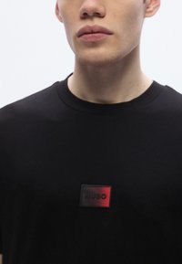 Μαύρο βαμβακερό μπλουζάκι με ορθογώνιο μπατζάκι λογότυπου με την επιγραφή "HUGO" σε κόκκινο. Στρογγυλή λαιμόκοψη και κοντά μανίκια, απαλής υφής.