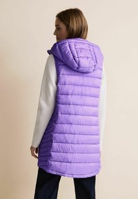 Lila puffy väst med huva, med horisontella kviltade linjer och en slät, glänsande yta. Bärs över en ljusfärgad långärmad topp.