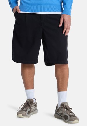 Schwarze Shorts mit Falten, knielang, aus Baumwolle. Getragen mit weißen Sportsocken und braunen Sportschuhen. Am oberen Rand ist ein blauer Pullover sichtbar.