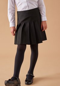 Schwarzer, gefalteter Rock mit einem taillierten Bund, kombiniert mit dunklen Strumpfhosen und schwarzen lackierten Schuhen mit einer Schleifenakzent. Weißes Hemd mit Knöpfen sichtbar.