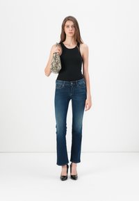 Svart tank topp, mørkeblå vide jeans og svarte spisse hæler. Holder en veske med slangeskinnsmønster. Modell med langt brunt hår og nøytral bakgrunn.