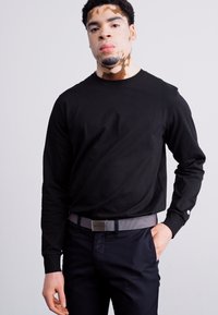 Fekete, hosszú ujjú póló kerek nyakkivágással, sötét nadrággal és mintás szürke övvel párosítva. Sima anyag, testhez álló fazon.