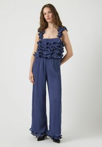 Navyblauwe jumpsuit met geruffelde, gelaagde schouderbanden en gestructureerde stof. Kenmerkt zich door wijde broekspijpen en geruffelde zoomdetails.