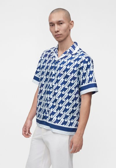 Hombre con una camisa de manga corta con botones y estampado azul y blanco, con pantalones blancos, de pie frente a un fondo liso y claro.