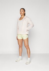 Chaqueta con cremallera rosa claro, camiseta beige y pantalones cortos amarillos claros. Zapatillas blancas con acentos en tonos pastel. Tejido suave y corte atlético.