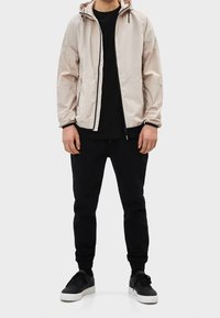 Ljusbeige zip-up-jacka med huva, elastiska muddar och en svart dragkedja, bärs över en svart tröja och svarta byxor. Svarta sneakers kompletterar looken.