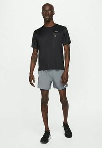 Μαύρο αθλητικό μπλουζάκι με κοντά μανίκια, που φέρει ένα μικρό λογότυπο, σε συνδυασμό με ανοιχτό γκρι σορτς. Φοριέται με μαύρα αθλητικά παπούτσια.