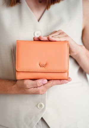 Peach-colored leather wallet with a folding design, featuring embossed logo and stitched edges. Held in hands against a light background.
