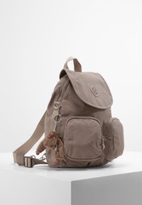 Khaki stof rygsæk med klaplukning, flere lynlåslommer og en blød abe nøglering. Har beige stropper og tekstureret overflade.