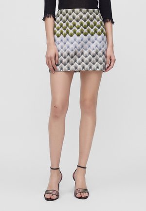 Femme portant une jupe courte aux motifs zigzag verts, gris et blancs et des sandales à talons hauts et bride à la cheville noires, debout devant un fond uni.