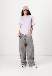 Tricou oversized lavandă cu grafică pe față, asortat cu blugi gri cu picioare largi, având un model decolorat și cusături în contrast. Include o șapcă bej.