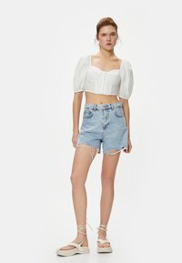Weißes Cropped-Top mit Puffärmel und strukturiertem Design, kombiniert mit hellblauen, hochtaillierten Jeansshorts mit fransigen Säumen. Getragen mit beigen Sandalen.