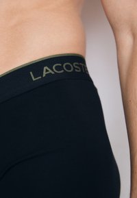 Close-up da cintura de uma pessoa a usar roupa interior Lacoste preta com uma faixa verde e o texto da marca Lacoste visível.