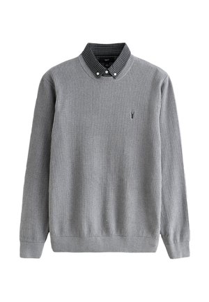 Pilkos spalvos megztinis su iškiliais briaunuotais raštais, apvalia iškirpte, ilgomis rankovėmis ir žvilgsnį traukiančiu juodo ir balto languoto marškinio apykakliu. Krūtinėje mažas logotipas.