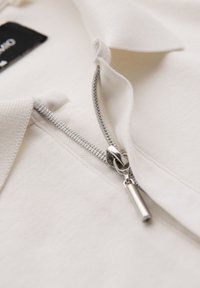 Krémová polo košile s polouzavřeným zipem se stříbrným kovovým táhlem a texturovaným detailem límce. Čistý, minimalistický design.