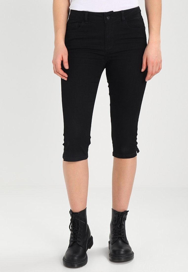 Femme portant un pantalon skinny noir raccourci et des bottines noires à lacets, debout devant un fond clair uni.