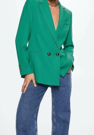 Femme portant un blazer vert croisé avec des boutons dorés et un jean bleu ample, une main dans la poche et l'autre reposant sur la cuisse.